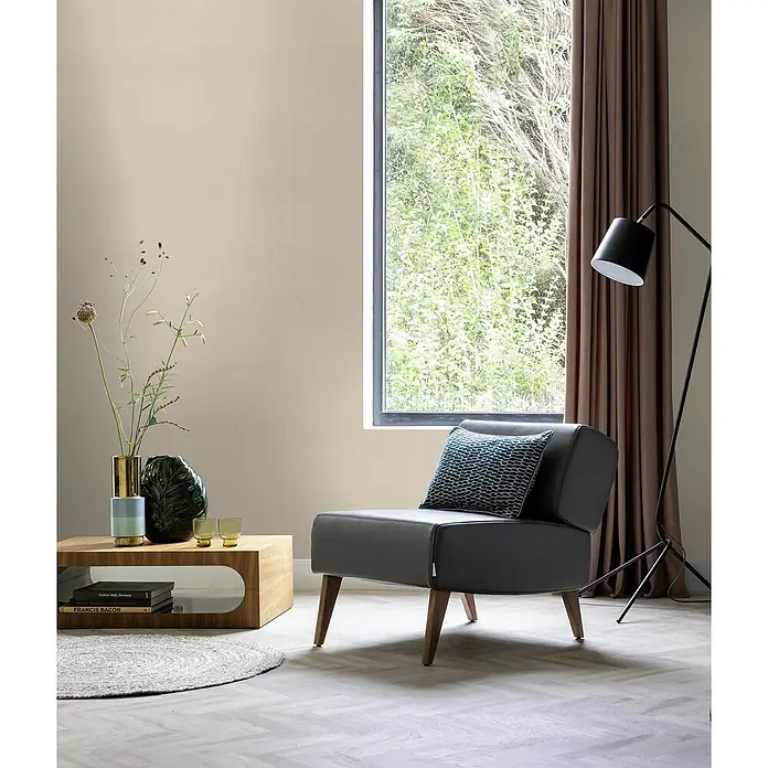 Rasch Selection Vliestapete Vinyl - Beigetaupe, Uni, 10,05 x 0,53 m - Das Bild zeigt einen grauen Sessel mit einem blauen Kissen in einem hellen Raum mit Holztisch und Fenster.