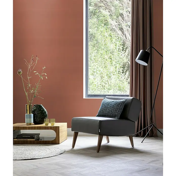 Rasch Selection Vliestapete Vinyl - Rotbraun, Uni, 10,05 x 0,53 m - Wohnzimmer mit modernem grauem Sessel mit blauem Kissen, Couchtisch aus Holz, Stehlampe und Fenster mit Blick auf Bäume.