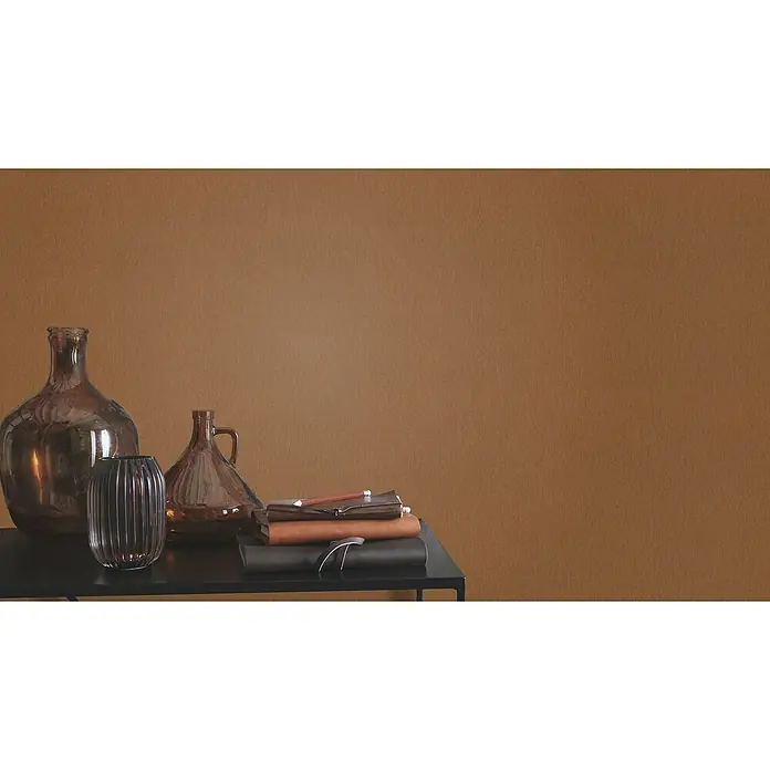 Rasch Incanto Vliestapete - Dunkelbraun, Uni, 10,05 x 0,53 m - Dekorative Stillleben-Arrangement mit Glaswaren und Notizbüchern auf einem eleganten schwarzen Tisch vor einem warmen, strukturierten Hintergrund.