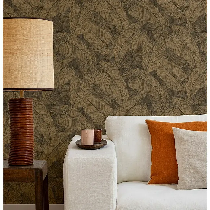 Rasch Amara Vliestapete Blätter - Kupferschwarz, Floral, 10,05 x 0,53 m - Brauner Tisch mit Lampe und weißes Sofa mit Kissen. Im Hintergrund eine Wand mit Blättertapete.