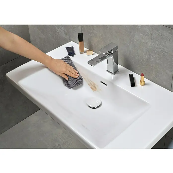 Villeroy & Boch Subway 3.0 Aufsatzwaschbecken - 80 x 47 cm, Mit schmutzabweisender Glasur, Mit Überlauf, Weiß Alpin, Anzahl Hahnlöcher: 1 - Weißes Keramikwaschbecken mit einer Hand, die einen Make-up-Fleck mit einem grauen Tuch reinigt. Kosmetika und ein Chromwasserhahn sind sichtbar.