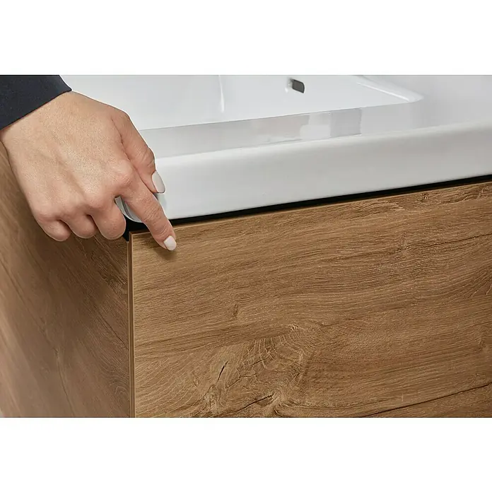 Villeroy & Boch Subway 3.0 Waschbeckenunterschrank - Kansas Oak, Anzahl Schubladen: 1 Stk., Farbe Griff: Volcano Black, B x T x H: 77,2 x 47,8 x 42,9 cm - Eine Hand berührt den grifflosen Holzunterschrank eines Badezimmermöbels mit weißem Waschbecken.