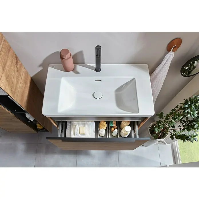 Villeroy & Boch Subway 3.0 Waschbeckenunterschrank - Kansas Oak, Anzahl Schubladen: 1 Stk., Farbe Griff: Volcano Black, B x T x H: 77,2 x 47,8 x 42,9 cm - Eine Aufsicht auf ein weißes Waschbecken mit matt-schwarzem Wasserhahn, integriert in einen Holzwaschtisch mit geöffneter, organisierter Schublade.