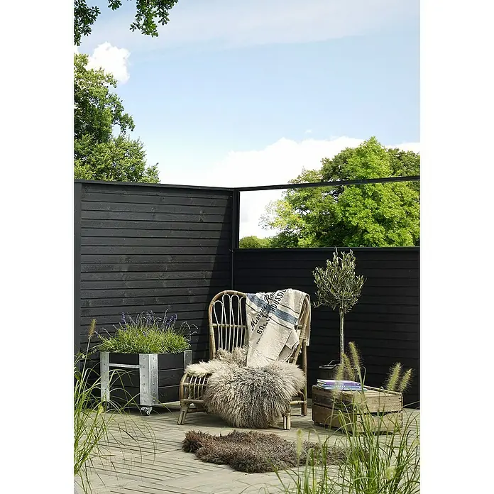 Plus Sichtschutzelement Plank - 174 x 129 cm, Schwarz (RAL 9005) - Einladender Außenbereich mit einem Rattansessel, einer Pflanze im Topf und einer Decke auf einer Holzterrasse vor einem schwarzen Zaun.