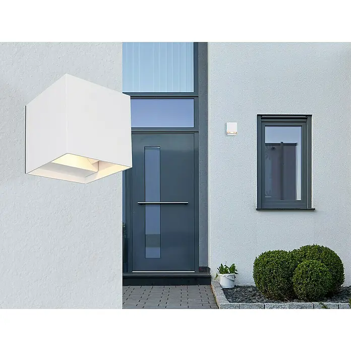 Globo LED-Außenwandleuchte Veronika - 3 W, 10 x 10 x 10 cm, Weiß, IP54 - Nahaufnahme einer modernen, würfelförmigen weißen Wandleuchte, die ein warmes Licht ausstrahlt. Sie ist an einer Fassade neben einer Haustür montiert.