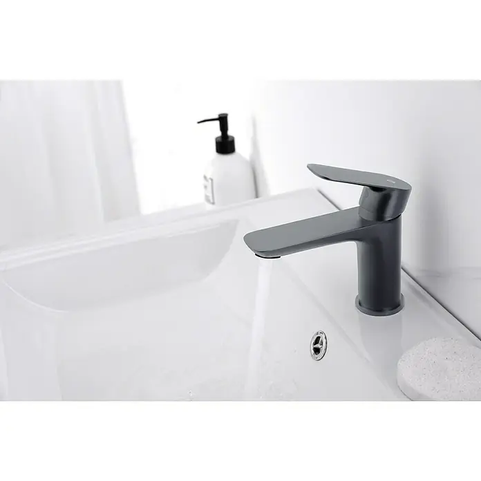 IO Cassio Grifo de lavabo - Titanio - Primer plano de un elegante grifo de baño negro mate sobre un lavabo de cerámica blanca con un dispensador de jabón al fondo.
