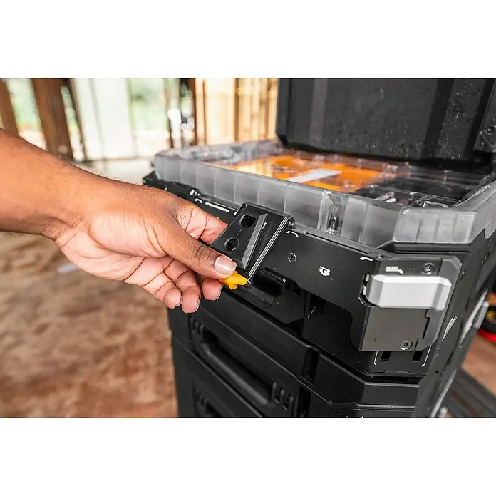 ToughBuilt StackTech Halterung - L x B x H: 40 x 5 x 8 cm, Schwarz - Nahaufnahme eines schwarzen Werkzeugkastens mit transparentem Deckel und gelbem Knopf, der von einer Hand bedient wird.\n