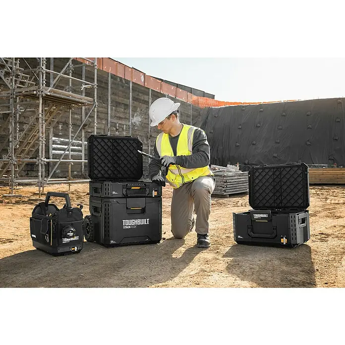 ToughBuilt StackTech Werkzeugkoffer-Set 3-tlg. - L x B x H: 50 x 56 x 83 cm, 3 -tlg., Räder - Ein Bauarbeiter kniet neben mehreren schwarzen Toughbuilt Stacktech Werkzeugkisten auf einer Baustelle.