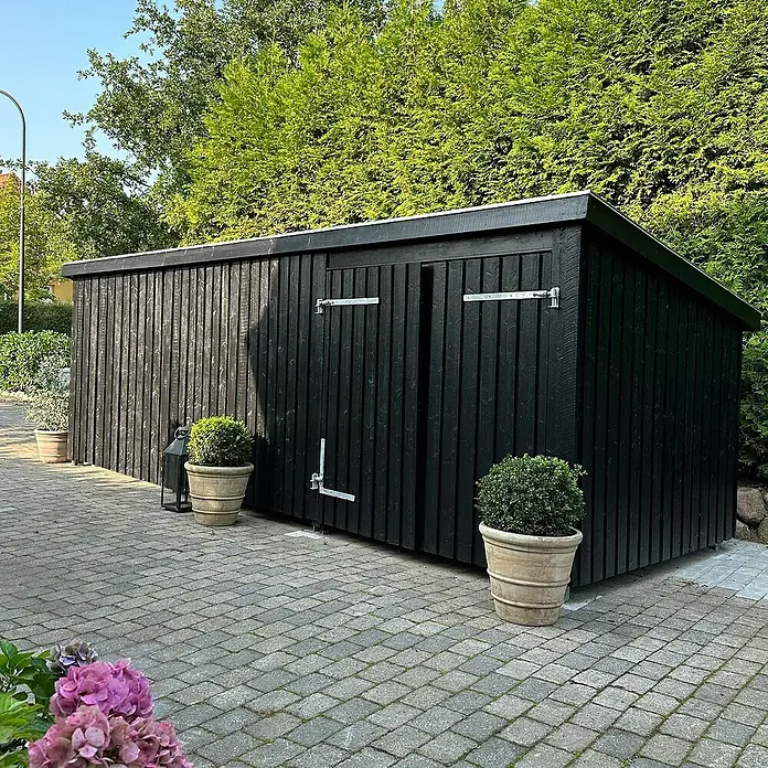 Plus Gartenhaus Multi Nordic - Außenmaß inkl. Dachüberstand (B x T): 650 x 240 cm, Holz, Geschlossen inkl. H-Pfostenanker, Dachpappe und Aluleisten - Langes, schwarzes Holz-Gartenhaus mit Pultdach auf gepflasterter Terrasse, umgeben von Topfpflanzen und Hecken.