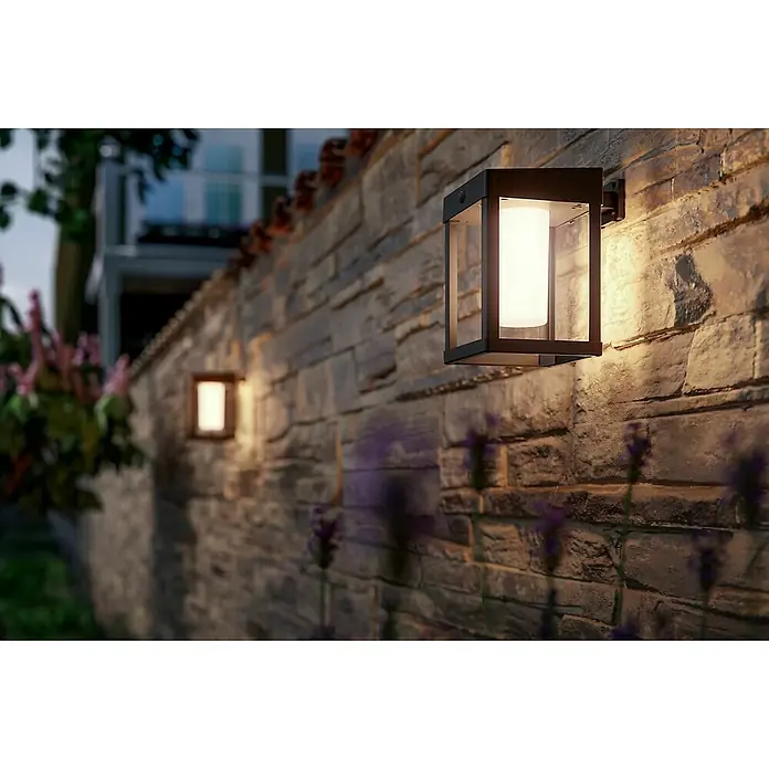 Philips Aplique solar LED para exterior Camill - 1,4 W, 19 x 14 x 20,5 cm, Negro, IP44, Blanco cálido - Luces de pared iluminan un muro de piedra, creando un ambiente cálido y acogedor en el exterior.