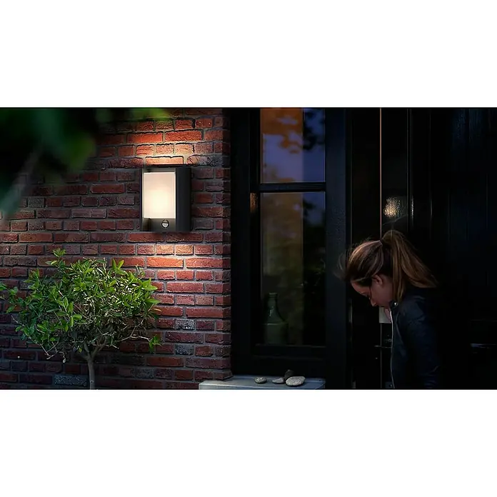 Philips Arbour LED-Sensor-Außenwandleuchte - 3,8 W, 16,5 x 7,5 x 22 cm, IP44 - Eine moderne Außenleuchte aus dunklem Metall mit Bewegungsmelder, die an einer roten Backsteinmauer neben einer Haustür angebracht ist.