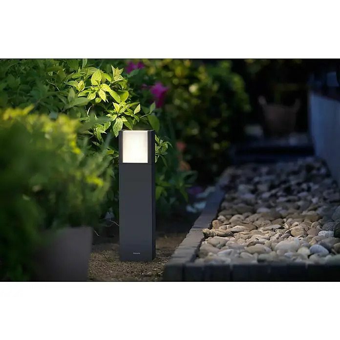 Philips Arbour Außensockelleuchte - Warmweiß, 10 x 10 x 40 cm, Mit Leuchtmittel - Aufnahme einer Philips Außenleuchte, die einen Gartenweg mit Pflanzen und Steinen beleuchtet, die eine stimmungsvolle Atmosphäre erzeugt.