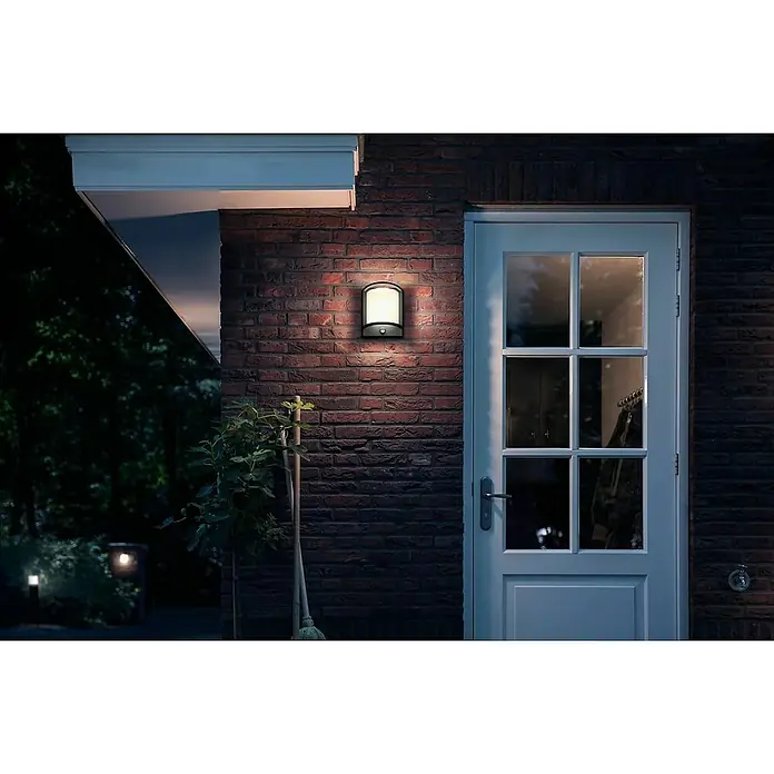 Philips LED-Sensor-Außenwandleuchte Samondra - 3,8 W, Lichtfarbe: Warmweiß, IP44 - Eine Außenwandleuchte mit Bewegungsmelder beleuchtet einen Backsteineingang neben einer weißen Tür in der Nacht.