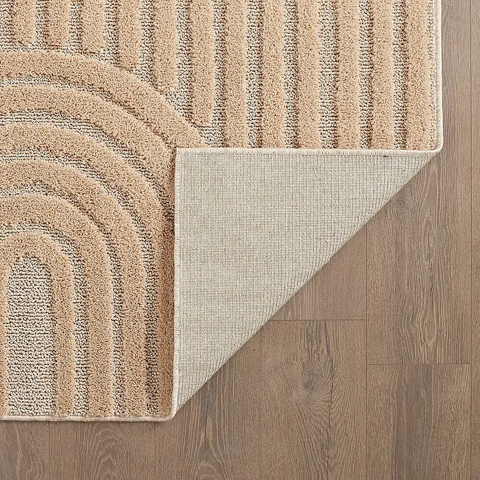 Kurzflorteppich Art - Rosa, 340 x 240 cm, 100 % Polypropylen - Nahaufnahme eines beigefarbenen Teppichs mit geometrischem Design, der auf einem Holzboden liegt und teilweise umgeschlagen ist, um die Unterseite zu zeigen.