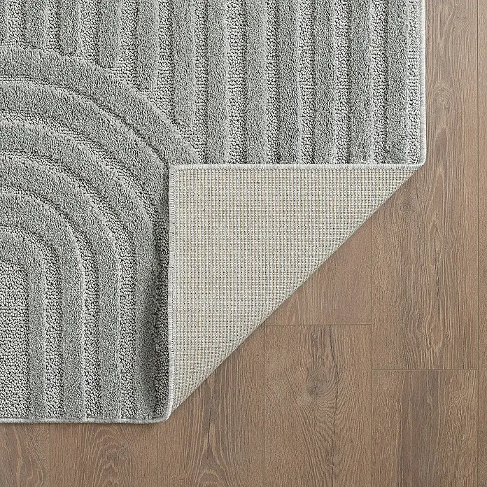 Kurzflorteppich Art - Silber, 290 x 200 cm, 100 % Polypropylen - Nahaufnahme eines grauen Badteppichs mit geometrischem Design auf Holzboden, der einen Teil der Unterseite des Teppichs zeigt.