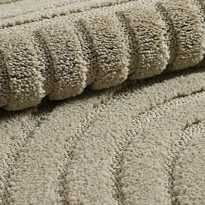 Kurzflorteppich Art - Beige, 150 x 80 cm, 100 % Polypropylen - Nahaufnahme eines gerollten, cremefarbenen Badteppichs mit gerippter Textur, der Komfort und Weichheit für das Badezimmer bietet.