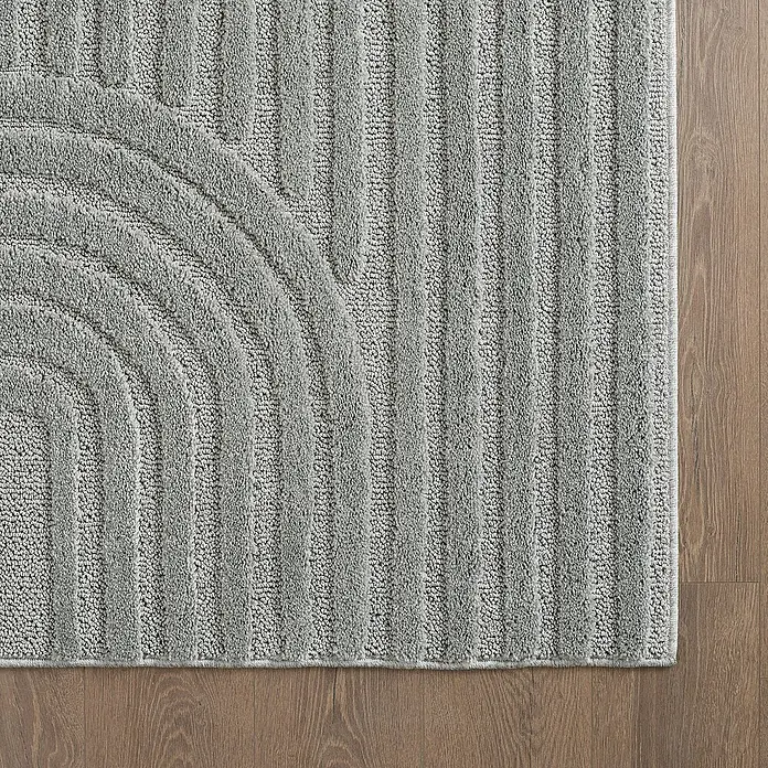 Kurzflorteppich Art - Silber, 290 x 200 cm, 100 % Polypropylen - Nahaufnahme einer grauen Badematte mit geometrischen Linien und Halbkreis auf einem Holzboden, die Textur und Design hervorhebt.