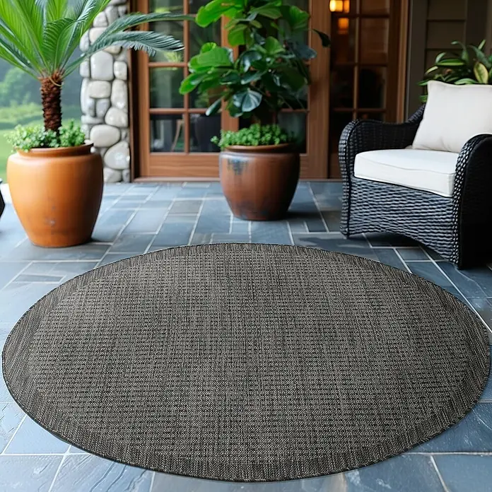Outdoor-Teppich Giza 1410 - Schwarz, Durchmesser: 200 cm, 100 % Polypropylen - Ein grauer, runder Teppich liegt auf einer Terrasse mit Pflanzen und einem Sessel. Der Teppich ist das Hauptmerkmal.