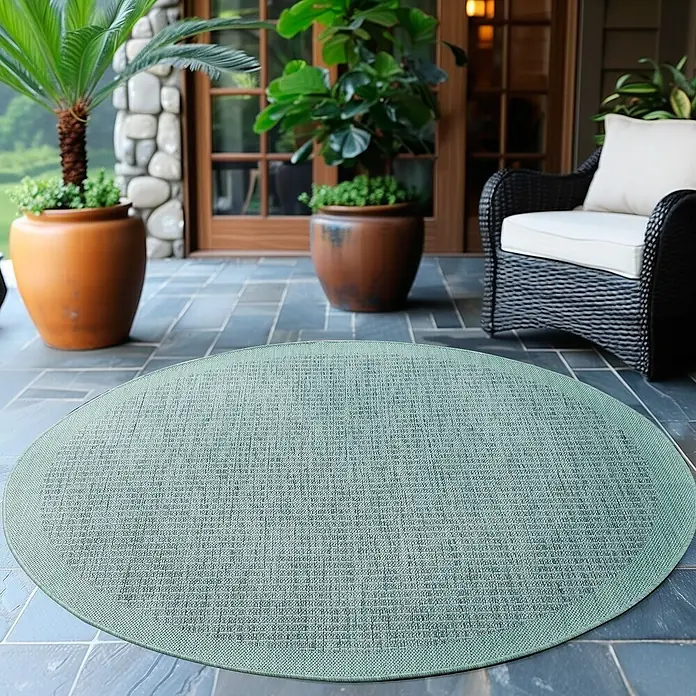 Outdoor-Teppich Giza 1410 - Blau, Durchmesser: 160 cm, 100 % Polypropylen - Ein hellgrüner, runder Teppich liegt auf einer Terrasse mit Pflanzen und einem Sessel im Hintergrund. Der Teppich ist der Blickfang.