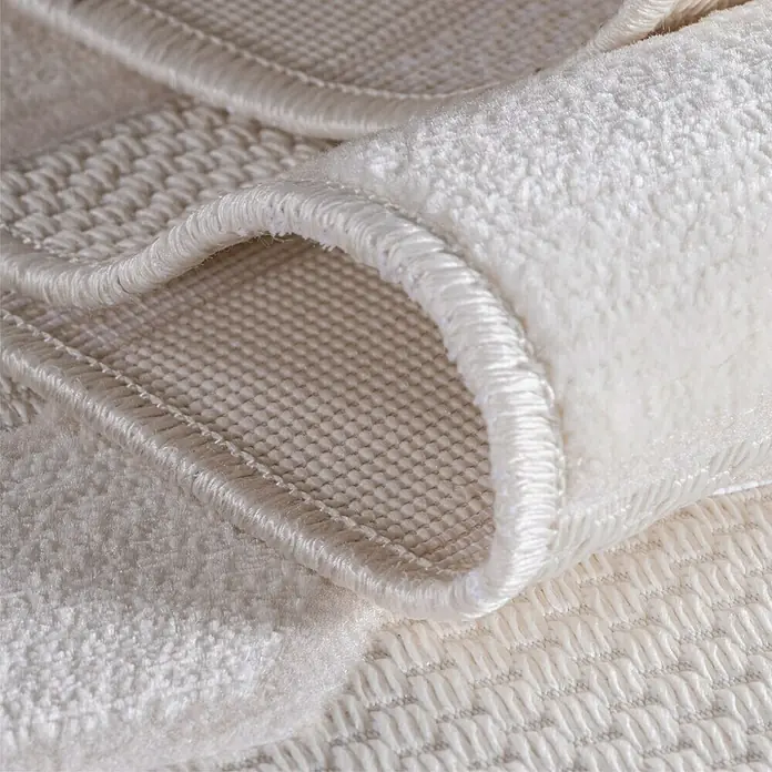 Kurzflorteppich Doha 1654 - Creme, 340 x 240 cm, 100 % Polypropylen - Nahaufnahme eines cremefarbenen Teppichs mit aufgerollter Kante, der die Textur und die hochwertige Verarbeitung hervorhebt.