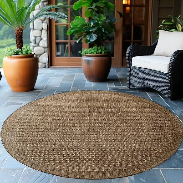 Outdoor-Teppich Giza 1410 - Braun, Durchmesser: 200 cm, 100 % Polypropylen - Ein brauner, ovaler Teppich liegt auf einer blauen Terrasse, umgeben von Pflanzen und einem Korbsessel. Der Teppich ist der Fokus.