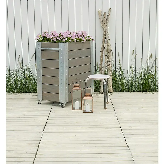 Plus Pflanzkasten Cubic - Außenmaß (L x B x H): 87 x 50 x 95 cm, Holz, Grau/Braun - Ein großer grauer Holzpflanzkübel mit Metallakzenten und Rollen, gefüllt mit rosa Blumen, auf einer hellen Terrasse.