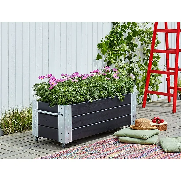 Plus Pflanzkasten Cubic - Außenmaß (L x B x H): 120 x 50 x 45 cm, Holz, Schwarz/Silber - Schwarzer Pflanzkasten aus Holz mit verzinkten Metallakzenten und Rollen, gefüllt mit grünen Pflanzen und rosa Blüten, auf Holzterrasse.