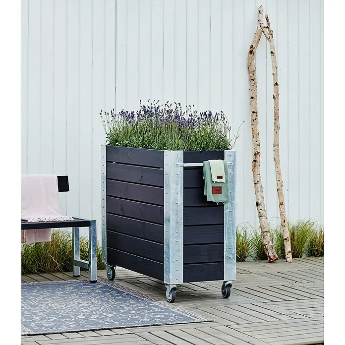 Plus Pflanzkasten Cubic - Außenmaß (L x B x H): 120 x 50 x 95 cm, Holz, Schwarz/Silber - Großer dunkler Holz-Pflanzkasten mit verzinkten Metallecken, Rollen, Lavendelpflanzen und einem Handtuch, auf einer Außenterrasse.