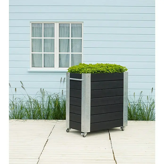 Plus Pflanzkasten Cubic - Außenmaß (L x B x H): 87 x 50 x 95 cm, Holz, Schwarz/Silber - Großer schwarzer Holzpflanzkübel mit Metallakzenten und grünen Pflanzen, auf Rollen, auf hellem Deck im Freien.