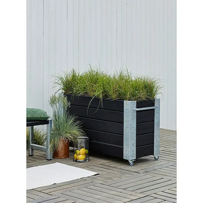 Plus Pflanzkasten Cubic - Außenmaß (L x B x H): 120 x 50 x 70 cm, Holz, Schwarz/Silber - Großer schwarzer Lattenholzpflanzkasten mit verzinkten Metallakzenten und Rollen, gefüllt mit grünem Gras auf einer Holzterrasse.