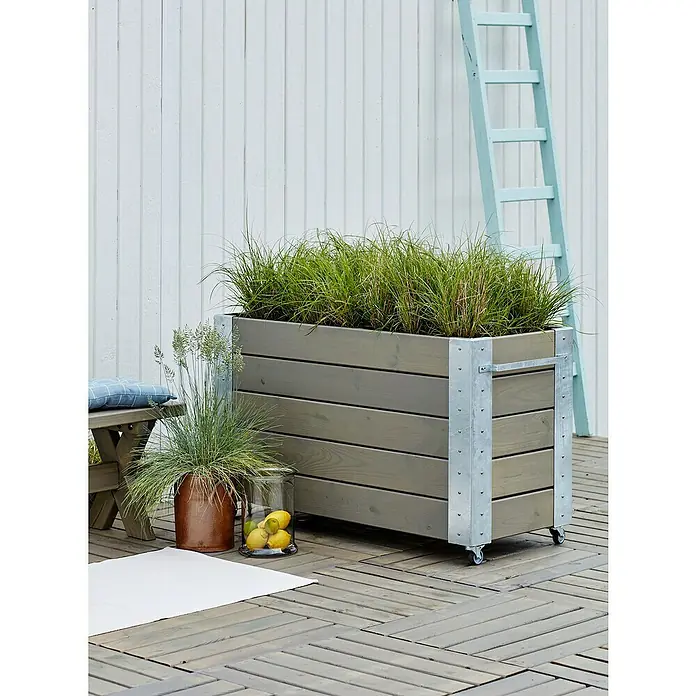 Plus Pflanzkasten Cubic - Außenmaß (L x B x H): 120 x 50 x 70 cm, Holz, Grau/Braun - Großer grauer Holzpflanzkasten auf Rollen, gefüllt mit grünem Gras, mit silbernen Metalleckhalterungen und Griffen, auf einer Holzterrasse.
