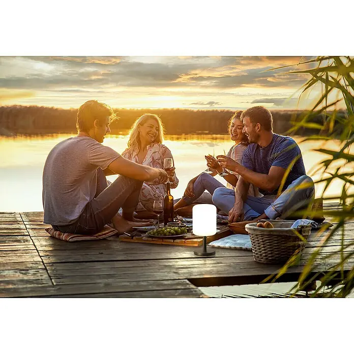 FHL easy Akku-Tischleuchte Volterra Pro - 2 W, L x B x H: 12 x 12 x 36 cm, Sand, RGBW - Weiße zylindrische Lampe mit dunklem Sockel, die ein Picknick auf einem Steg bei Sonnenuntergang beleuchtet.