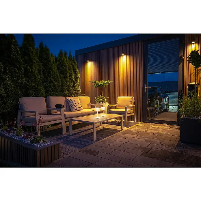 FHL easy Akku-Tischleuchte Finley - 1 W, L x B x H: 8,7 x 8,7 x 22 cm, Sand, Warmweiß - Elegante Outdoor-Lounge-Möbel mit hellen Kissen und zwei leuchtenden weißen Tischlampen auf einer Terrasse, die eine gemütliche Abendatmosphäre schaffen.
