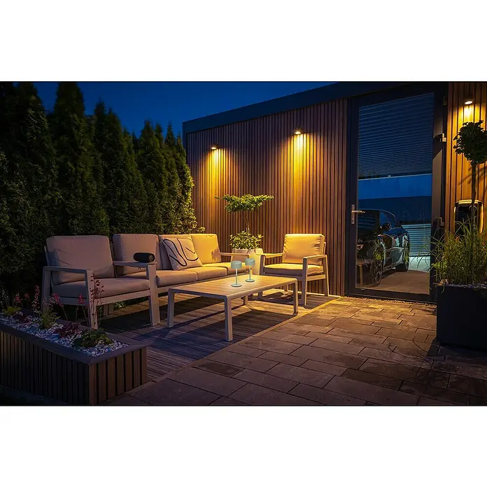 FHL easy Akku-Tischleuchte Finley - 1 W, L x B x H: 8,7 x 8,7 x 22 cm, Salbei, Warmweiß - Zwei kleine, hellgraue tragbare Tischlampen beleuchten einen Outdoor-Couchtisch auf einer Terrasse bei Dämmerung.