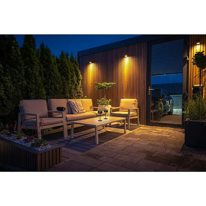 FHL easy Akku-Tischleuchte Finley - 1 W, L x B x H: 8,7 x 8,7 x 22 cm, Grau, Warmweiß - Zwei schwarze minimalistische Tischlampen beleuchten einen Outdoor-Couchtisch neben modernen Sitzgelegenheiten bei Dämmerung.