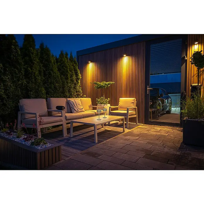 FHL easy Akku-Tischleuchte Finley - 1 W, L x B x H: 8,7 x 8,7 x 22 cm, Greige, Warmweiß - Eine Outdoor-Lounge-Sitzgruppe mit beigen Kissen und Couchtisch mit zwei Lampen, nachts auf einer Holzterrasse beleuchtet.