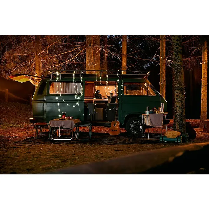 FHL easy LED-Lichterkette Benni - Innen, Lichtfarbe: Warmweiß, Akkubetrieben - Dunkelgrüner Camper Van mit Lichterkette, nachts im Wald geparkt, umgeben von Campingausrüstung.