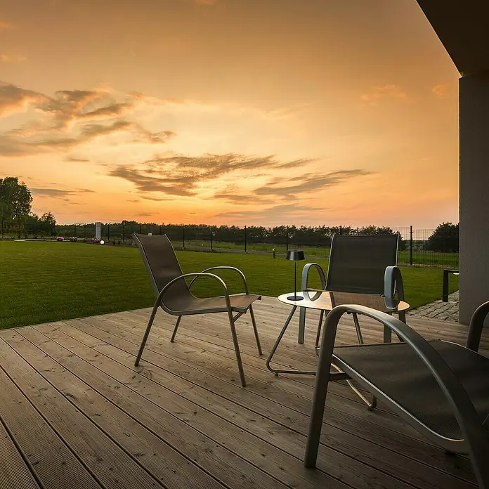 FHL easy Akku-Tischleuchte Verona - 1,5 W, L x B x H: 30 x 11 x 30 cm, Sandschwarz, Warmweiß - Gartenmöbel auf einer Holzterrasse bei Sonnenuntergang, mit dunklen Stühlen und einem kleinen Tisch mit Lampe.