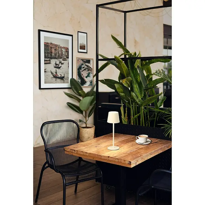 FHL easy Akku-Tischleuchte Roxi - 1,5 W, L x B x H: 9 x 9 x 27,5 cm, Sand, Warmweiß - Beige Tischlampe auf einem Holztisch, spendet Umgebungslicht in einer Café-Umgebung.