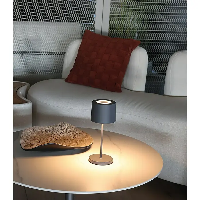 FHL easy Akku-Tischleuchte Bologna Pro - 2 W, L x B x H: 12 x 12 x 28 cm, Sand, RGBW - Eine dunkelgraue, zylindrische, tragbare Tischlampe auf einem weißen Marmortisch spendet warmes Umgebungslicht in einer Lounge.