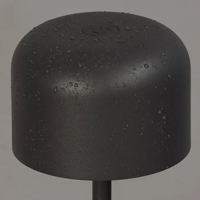 FHL easy Akku-Tischleuchte Finley - 1 W, L x B x H: 8,7 x 8,7 x 22 cm, Sandgrau, Warmweiß - Strukturiertes dunkelgraues, kuppelförmiges Gerät mit Einschaltsymbol, bedeckt mit Wassertropfen, auf einem dunklen Ständer.