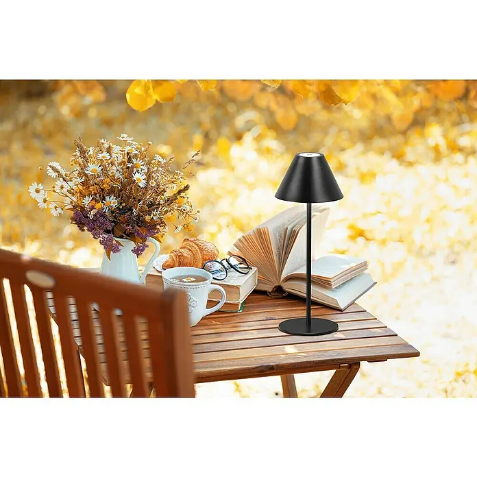 FHL easy Akku-Tischleuchte Kano - 2 W, L x B x H: 14 x 14 x 36 cm, Sandschwarz, Warmweiß - Schwarze Tischlampe auf einem Holztisch in herbstlicher Outdoor-Umgebung, beleuchtet Bücher.