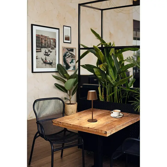 FHL easy Akku-Tischleuchte Roxi - 1,5 W, L x B x H: 9 x 9 x 27,5 cm, Rost, Warmweiß - Braune kabellose Tischlampe auf einem Holztisch im Café neben einer Kaffeetasse, spendet stimmungsvolles Licht.