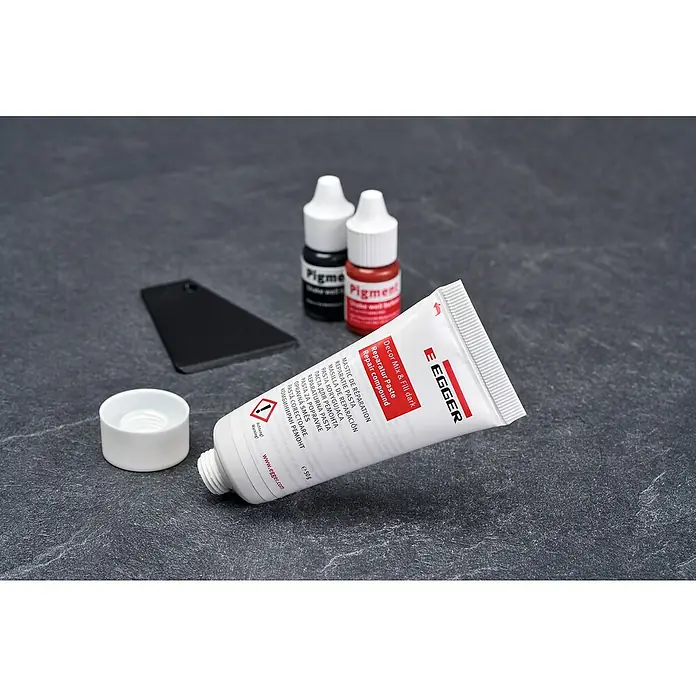 Egger Reparatur-Set Mix & Fill Dark Decor - Farbgebung: Dunkel, 5 -tlg. - EGGER Reparaturpaste-Tube mit Kappe, zwei Pigmentflaschen (schwarz, rot) und ein schwarzes Applikationswerkzeug auf dunkler Oberfläche.