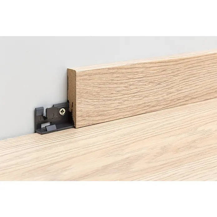 Egger Leistenclip - 30 Stk. - Nahaufnahme einer Holzfußleiste mit Befestigungsclip an einer Wand. Die Fußleiste ist hellbraun und hat eine Holzmaserung.