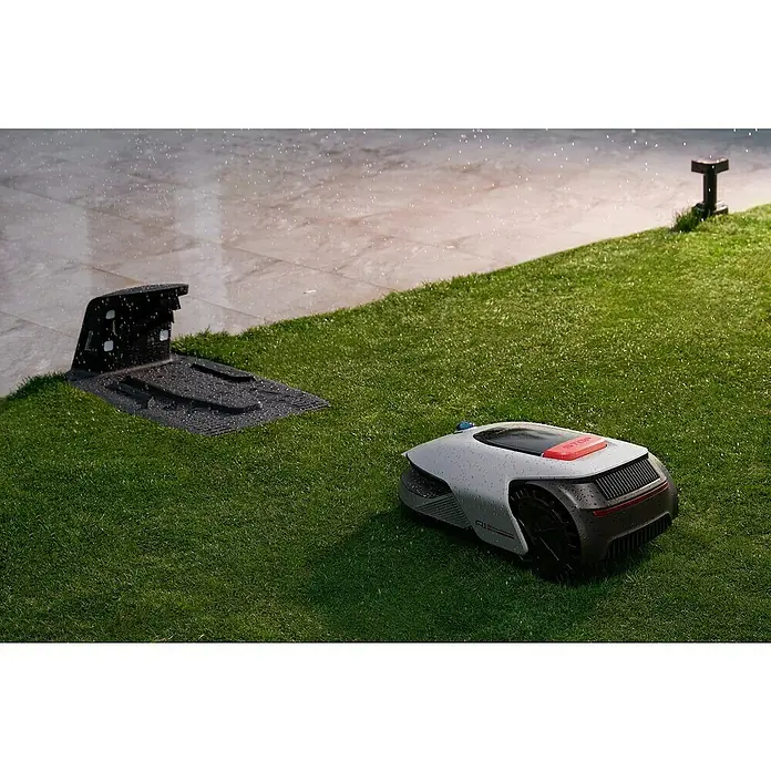 Dreame Mähroboter A1 Pro - 2.000 m², Schnittbreite: 22 cm, Laser-gestützte Navigation, LiDAR - Ein Rasenmähroboter steht im Regen neben seiner Ladestation auf einer grünen Rasenfläche.