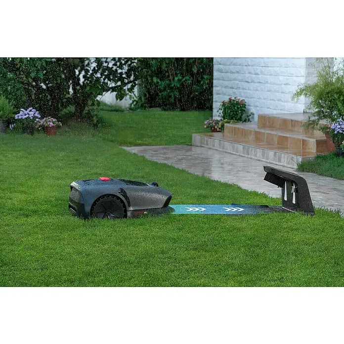 Mova Mähroboter 600 Plus - 600 m², Schnittbreite: 20 cm, Laser-gestützte Navigation, LiDAR - Ein Rasenmähroboter fährt im Regen auf einer Ladeplattform. Das Gerät ist schwarz und hat einen roten Knopf. Das Gras ist saftig grün.