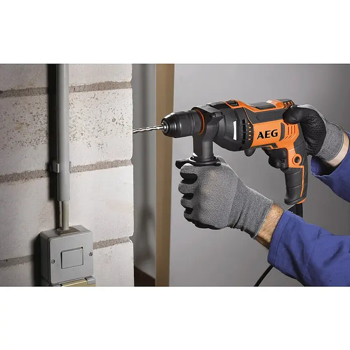 AEG Schlagbohrmaschine SBE 750 RE - 750 W, Schnellspannbohrfutter - Behandschuhte Person bohrt mit einem orange-schwarzen AEG Bohrhammer in eine helle Ziegelwand neben einem Elektrokabelkanal.