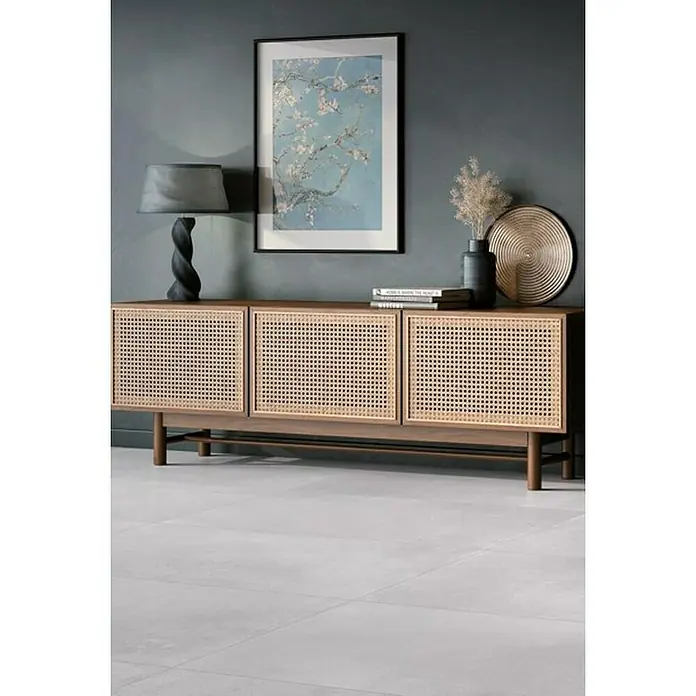 Pavimento porcelánico Mood - 60,5 x 60,5 cm, Pearl, Mate - Un elegante aparador de madera con frentes de ratán, decorado con una lámpara, libros y objetos decorativos encima.