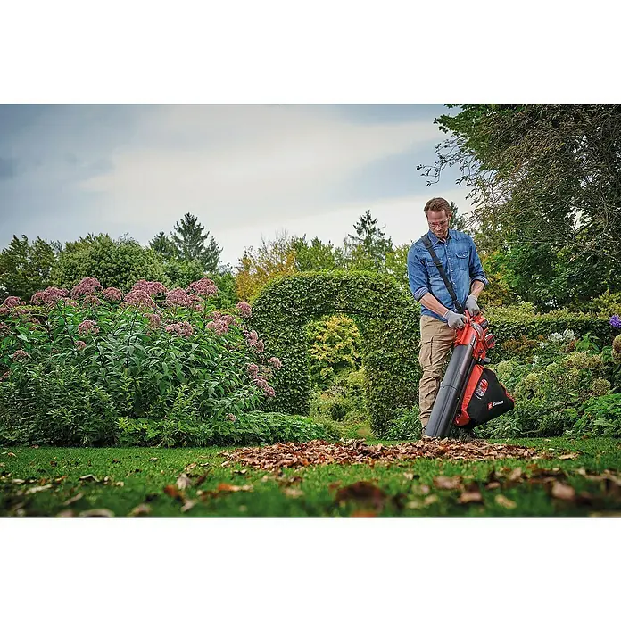 Einhell Professional Akku-Laubsauger & -Laubbläser VENTURRO 36/240 - 18 V, Ohne Akku, Blasgeschwindigkeit: 240 km/h - Ein Mann benutzt einen Einhell-Laubsauger im Garten, um Herbstlaub zu entfernen. Das Gerät ist rot und schwarz und mit einem Tragegurt ausgestattet.